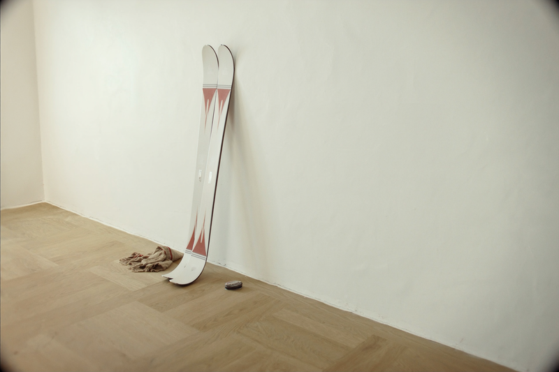 Paire de skis rangée debout contre un mur blanc, avec un chiffon et un fart posés au sol