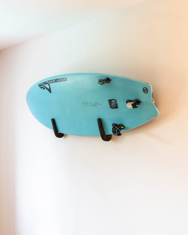 Black-tinted solid oak surfboard wall rack with a board displayed flat on the wall
